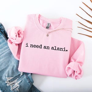 Op de afbeelding: Een lichtroze sweatshirt met de tekst "i need an alani" in het zwart. Het sweatshirt wordt gecombineerd met een blauwe spijkerbroek en een delicate ketting. Op de achtergrond een palmblad.