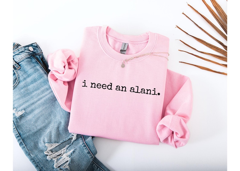Op de afbeelding: Een lichtroze sweatshirt met de tekst "i need an alani" in het zwart. Het sweatshirt wordt gecombineerd met een blauwe spijkerbroek en een delicate ketting. Op de achtergrond een palmblad.
