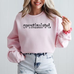 Puede incluir: Una sudadera rosa claro con la palabra "overstimulated." impresa en negro, con fuente de m&aacute;quina de escribir. La parte superior de manga larga se combina con pantalones cortos de mezclilla descoloridos. Una scrunchie rosa es visible en la mu&ntilde;eca.