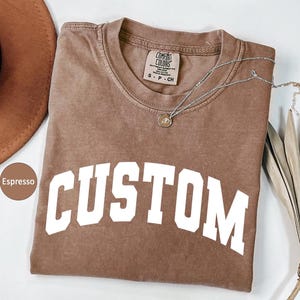 May include: Espresso-colored t-shirt with the word "CUSTOM" in white block letters. The shirt has a crew neck and a small tag at the neckline. A silver necklace with a pendant is draped across the shirt.