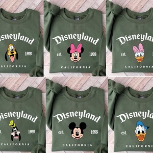 Puede incluir: Sudaderas verde oliva con la palabra "Disneyland" en escritura blanca, con varios rostros de personajes de Disney. Las sudaderas tambi&eacute;n muestran "est. 1955" y "CALIFORNIA" en texto blanco. Los personajes incluyen a Goofy, Minnie Mouse y Donald Duck.