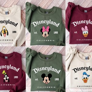 Puede incluir: Una colecci&oacute;n de sudaderas en varios colores, cada una con un dise&ntilde;o de Disneyland. Los dise&ntilde;os incluyen a Pluto, Minnie Mouse, Daisy Duck, Goofy, Mickey Mouse y Donald Duck. El texto incluye "Disneyland", "est. 1955" y "California".