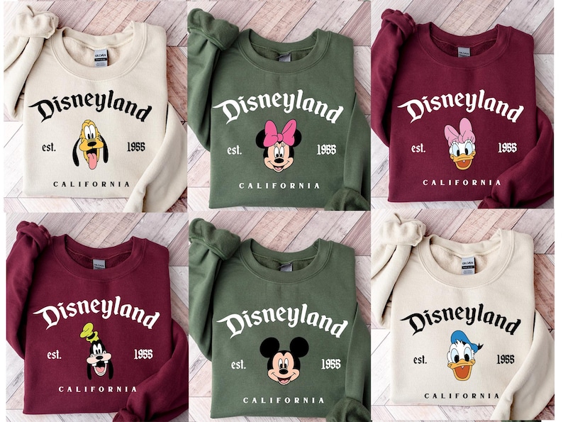 Puede incluir: Una colecci&oacute;n de sudaderas en varios colores, cada una con un dise&ntilde;o de Disneyland. Los dise&ntilde;os incluyen a Pluto, Minnie Mouse, Daisy Duck, Goofy, Mickey Mouse y Donald Duck. El texto incluye "Disneyland", "est. 1955" y "California".