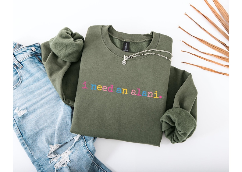 Op de afbeelding: Een olijfgroene sweatshirt met de geborduurde tekst "i need an alani" in kleurrijke letters. Een zilveren ketting ligt op de sweatshirt, die op een spijkerbroek ligt.