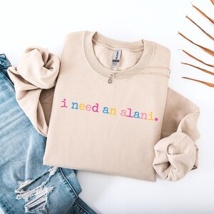 Op de afbeelding: Een beige sweatshirt met de zin "i need an alani." in regenboogkleurige letters. De sweatshirt wordt gecombineerd met lichtblauwe jeans met distressed details. Een ketting met een kleine hanger ligt op de sweatshirt.