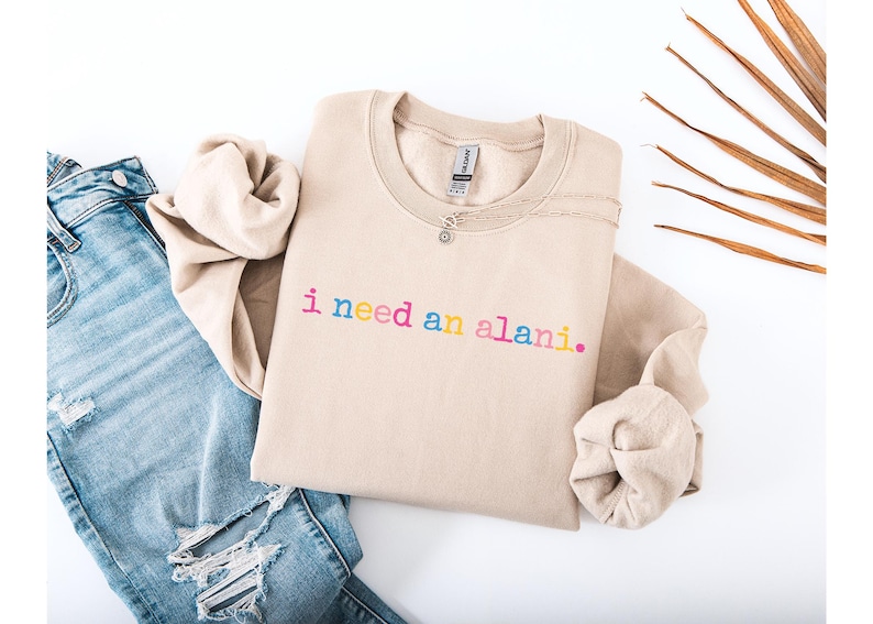 Op de afbeelding: Een beige sweatshirt met de zin "i need an alani." in regenboogkleurige letters. De sweatshirt wordt gecombineerd met lichtblauwe jeans met distressed details. Een ketting met een kleine hanger ligt op de sweatshirt.