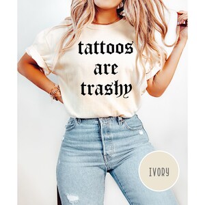 May include: Ivory-colored t-shirt with the text "tattoos are trashy" in a gothic font. The shirt is paired with light blue jeans. A round ivory-colored tag with the word "IVORY" is visible.