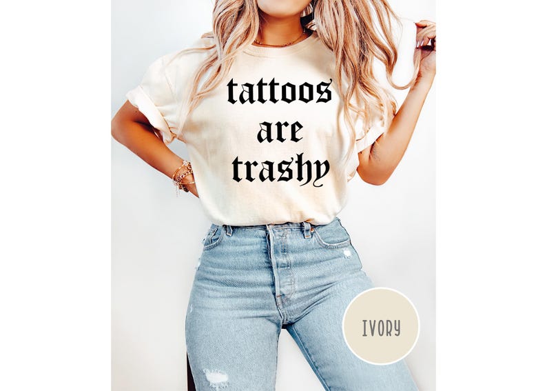 May include: Ivory-colored t-shirt with the text "tattoos are trashy" in a gothic font. The shirt is paired with light blue jeans. A round ivory-colored tag with the word "IVORY" is visible.