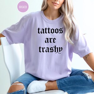 May include: A light purple t-shirt with the text "tattoos are trashy" in black gothic font. The shirt is worn with blue jeans. A small, round, pink sticker with the word "ORCHID" is in the upper left corner.