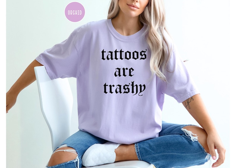 May include: A light purple t-shirt with the text "tattoos are trashy" in black gothic font. The shirt is worn with blue jeans. A small, round, pink sticker with the word "ORCHID" is in the upper left corner.