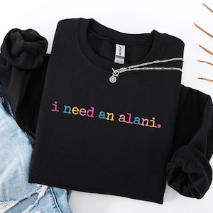Op de afbeelding: Zwart sweatshirt met ronde hals met de zin "i need an alani." geborduurd in regenboogkleuren. Een zilveren ketting met een ronde hanger is over het sweatshirt gedrapeerd. Het sweatshirt is op een wit oppervlak gevouwen.