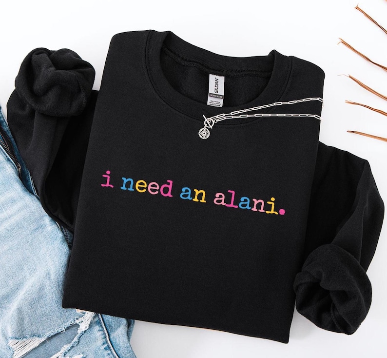 Op de afbeelding: Zwart sweatshirt met ronde hals met de zin "i need an alani." geborduurd in regenboogkleuren. Een zilveren ketting met een ronde hanger is over het sweatshirt gedrapeerd. Het sweatshirt is op een wit oppervlak gevouwen.