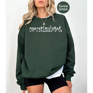 Puede incluir: Sudadera verde bosque con la palabra "overstimulated." en blanco, cuello redondo y mangas largas. La sudadera se combina con pantalones cortos negros y una camiseta interior blanca. El texto "Forest Green" est&aacute; en un c&iacute;rculo verde en la esquina superior derecha.