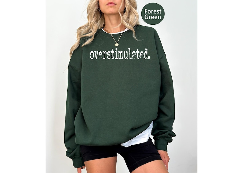 Puede incluir: Sudadera verde bosque con la palabra "overstimulated." en blanco, cuello redondo y mangas largas. La sudadera se combina con pantalones cortos negros y una camiseta interior blanca. El texto "Forest Green" est&aacute; en un c&iacute;rculo verde en la esquina superior derecha.
