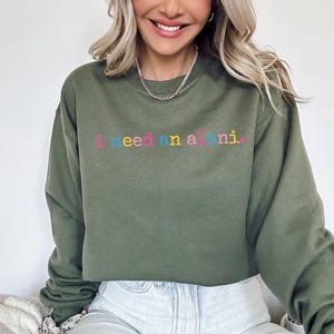 Op de afbeelding: Olijfgroene sweatshirt met de geborduurde tekst "I need an all nighter" in kleurrijke letters. De sweatshirt heeft een ronde hals en lijkt gemaakt van een zacht materiaal. De stijl is casual en comfortabel.
