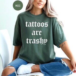 May include: Moss green t-shirt with the phrase "tattoos are trashy" in white gothic font. The shirt is a relaxed fit and is paired with blue jeans. A round moss-colored label is in the upper left corner.