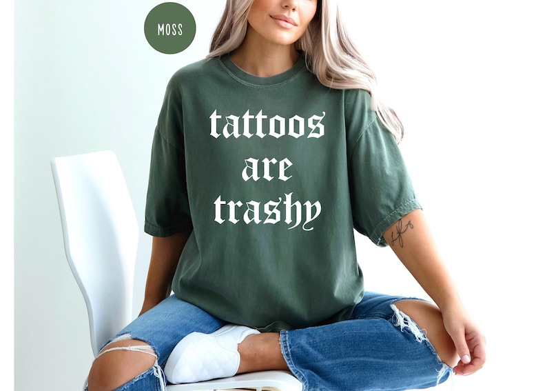 May include: Moss green t-shirt with the phrase "tattoos are trashy" in white gothic font. The shirt is a relaxed fit and is paired with blue jeans. A round moss-colored label is in the upper left corner.