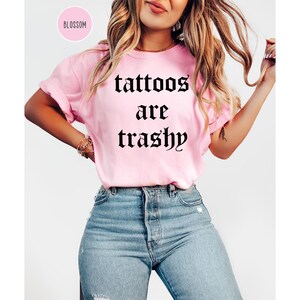 May include: A light pink t-shirt with the phrase "tattoos are trashy" in black gothic font. The shirt is paired with blue jeans. The word "Blossom" is in a pink circle in the upper left corner.