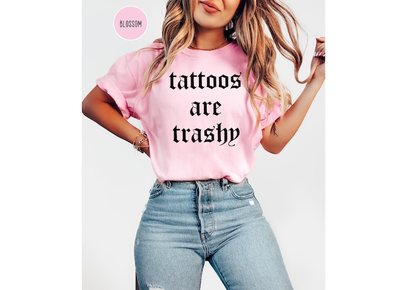 May include: A light pink t-shirt with the phrase "tattoos are trashy" in black gothic font. The shirt is paired with blue jeans. The word "Blossom" is in a pink circle in the upper left corner.