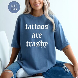 May include: Blue jean-colored t-shirt with the text "tattoos are trashy" in white gothic font. The shirt is a short-sleeved, oversized fit. A circular "Blue Jean" label is in the upper left corner.