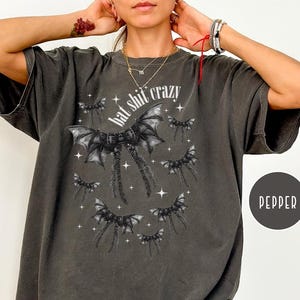 May include: Dark gray t-shirt with a bat-wing graphic and the text "hat shit crazy" in white. The design features multiple bat wings and stars. The shirt is a relaxed fit.