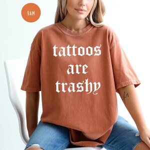 May include: A rust-colored t-shirt with the phrase "tattoos are trashy" in white gothic lettering. The shirt is a short-sleeved, oversized fit. The model is wearing blue jeans. A small orange circle with the letters "YAM" is in the upper left corner.