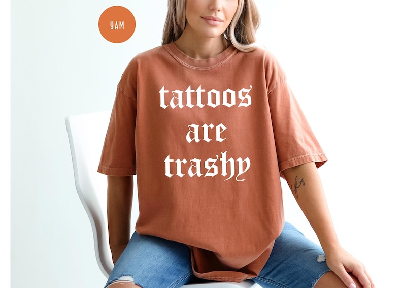 May include: A rust-colored t-shirt with the phrase "tattoos are trashy" in white gothic lettering. The shirt is a short-sleeved, oversized fit. The model is wearing blue jeans. A small orange circle with the letters "YAM" is in the upper left corner.