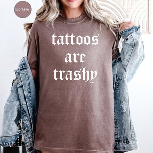 May include: Espresso-colored t-shirt with the text "tattoos are trashy" in a gothic font. The shirt is worn with a denim jacket. The shirt is a solid color and has a vintage look.