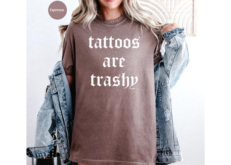 May include: Espresso-colored t-shirt with the text "tattoos are trashy" in a gothic font. The shirt is worn with a denim jacket. The shirt is a solid color and has a vintage look.