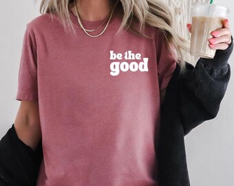 May include: A short-sleeved, mauve-colored t-shirt with the phrase "be the good" in white, bold, rounded letters. The shirt is worn with a black jacket. A clear plastic cup with a green straw and iced beverage is held in the hand.