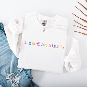 Op de afbeelding: Een witte sweatshirt met de tekst "i need an alani." in regenboogkleuren. De afbeelding toont een versleten blauwe jeans en een zilveren ketting. Een casual, alledaagse stijl.