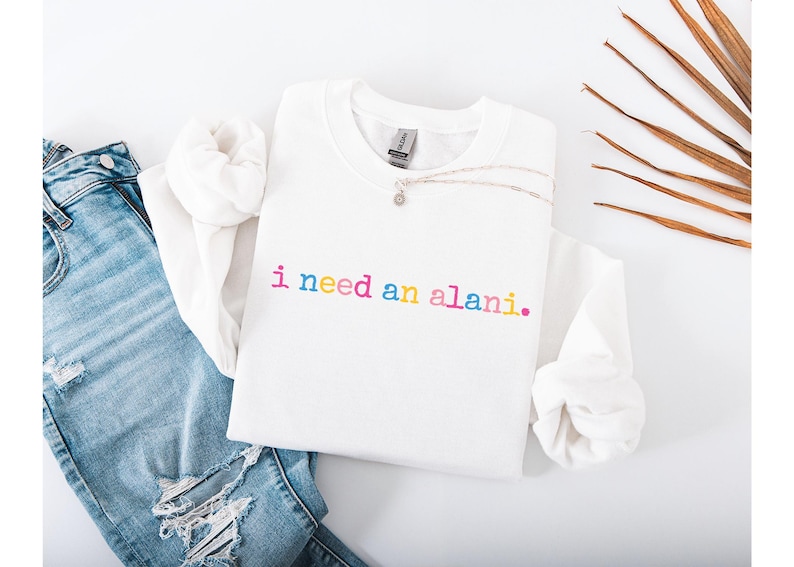 Op de afbeelding: Een witte sweatshirt met de tekst "i need an alani." in regenboogkleuren. De afbeelding toont een versleten blauwe jeans en een zilveren ketting. Een casual, alledaagse stijl.