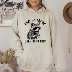 May include: Cream-colored hoodie with a black and white raccoon graphic holding a beverage. The text "HANG ON, LET ME OVERTHINK THIS" is printed above and below the raccoon illustration. The hoodie has a front pocket and a hood.