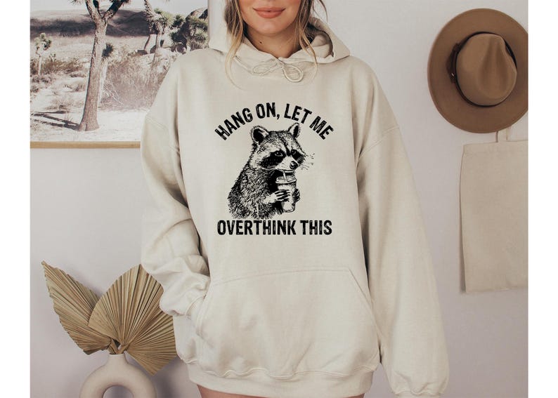 May include: Cream-colored hoodie with a black and white raccoon graphic holding a beverage. The text "HANG ON, LET ME OVERTHINK THIS" is printed above and below the raccoon illustration. The hoodie has a front pocket and a hood.