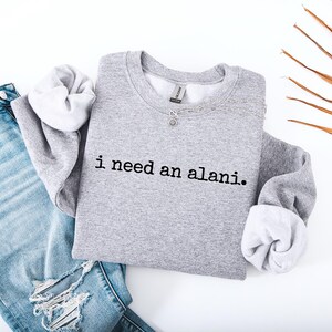 Op de afbeelding: Een grijze sweatshirt met de tekst "i need an alani." wordt getoond op een witte ondergrond naast een lichtblauwe ripped jeans. De sweatshirt heeft witte manchetten en een ronde hals. Een zilveren ketting is zichtbaar.