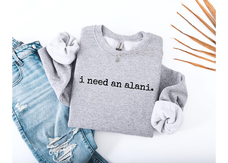 Op de afbeelding: Een grijze sweatshirt met de tekst "i need an alani." wordt getoond op een witte ondergrond naast een lichtblauwe ripped jeans. De sweatshirt heeft witte manchetten en een ronde hals. Een zilveren ketting is zichtbaar.