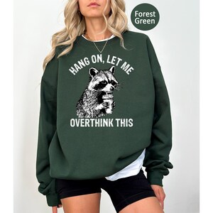 May include: Forest green sweatshirt with a raccoon graphic holding a drink. The text "HANG ON, LET ME OVERTHINK THIS" is above and below the raccoon illustration. The sweatshirt is a solid color.