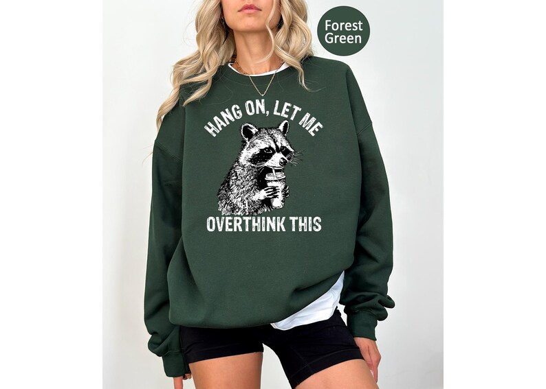 May include: Forest green sweatshirt with a raccoon graphic holding a drink. The text "HANG ON, LET ME OVERTHINK THIS" is above and below the raccoon illustration. The sweatshirt is a solid color.