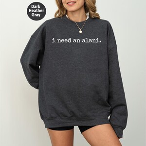 Op de afbeelding: Donkergrijs sweatshirt met de witte tekst "i need an alani.". De crew neck sweatshirt heeft lange mouwen. Een kleine, cirkelvormige afbeelding in de afbeelding geeft de kleur aan als "Dark Heather Gray".