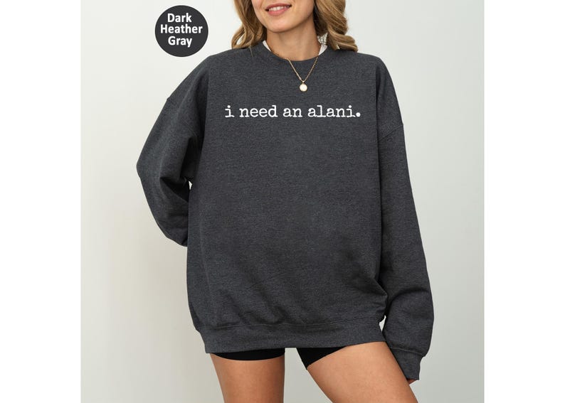 Op de afbeelding: Donkergrijs sweatshirt met de witte tekst "i need an alani.". De crew neck sweatshirt heeft lange mouwen. Een kleine, cirkelvormige afbeelding in de afbeelding geeft de kleur aan als "Dark Heather Gray".