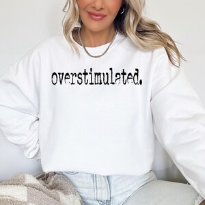 Puede incluir: Sudadera blanca con la palabra "overstimulated." impresa en negro, con fuente de m&aacute;quina de escribir. La parte superior de manga larga es de estilo casual, adecuada para el uso diario. La imagen muestra a una persona usando la sudadera.