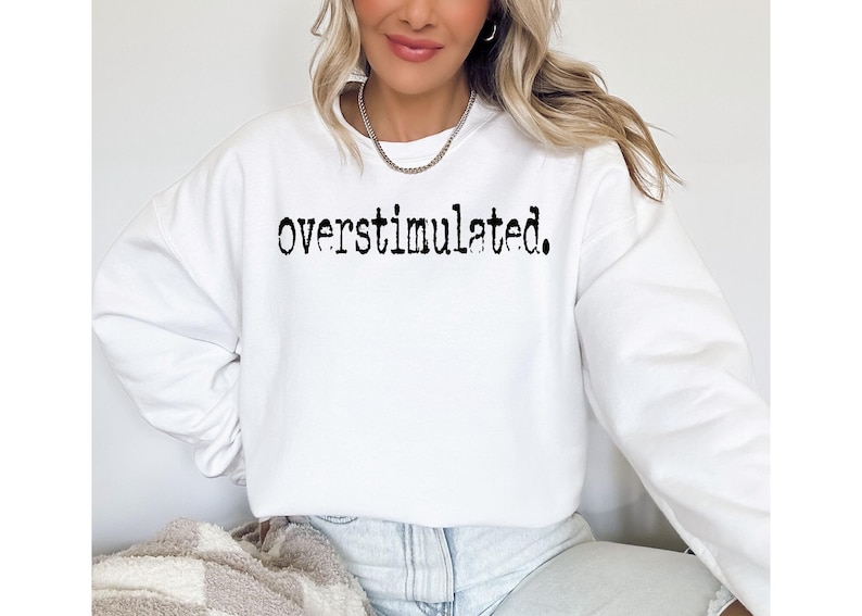 Puede incluir: Sudadera blanca con la palabra "overstimulated." impresa en negro, con fuente de m&aacute;quina de escribir. La parte superior de manga larga es de estilo casual, adecuada para el uso diario. La imagen muestra a una persona usando la sudadera.
