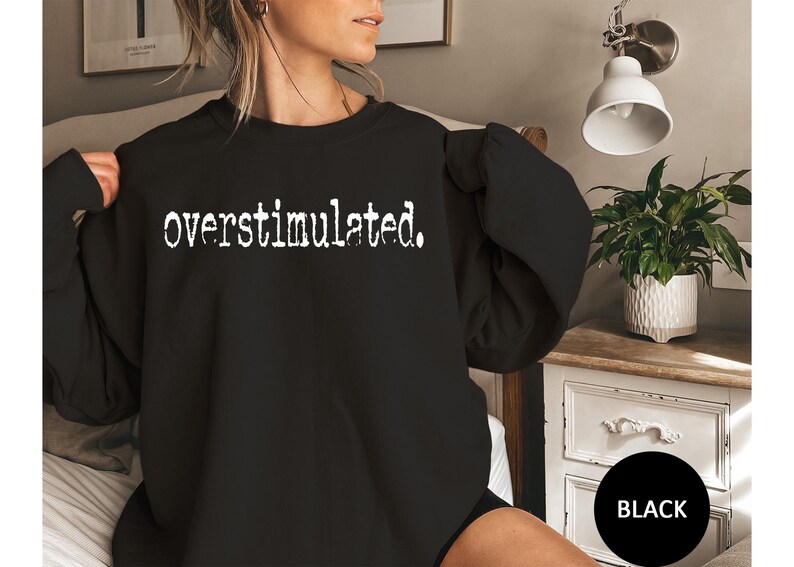 Puede incluir: Sudadera negra con la palabra "overstimulated" impresa en blanco con una fuente audaz. La sudadera es de color negro liso. La imagen tambi&eacute;n muestra una peque&ntilde;a mesita de noche blanca con una planta en una maceta blanca.