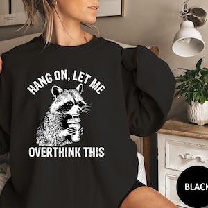 May include: Black sweatshirt featuring a raccoon holding a beverage, with the text "HANG ON, LET ME OVERTHINK THIS" in white. The raccoon graphic is detailed in black and white. The sweatshirt is a casual style.
