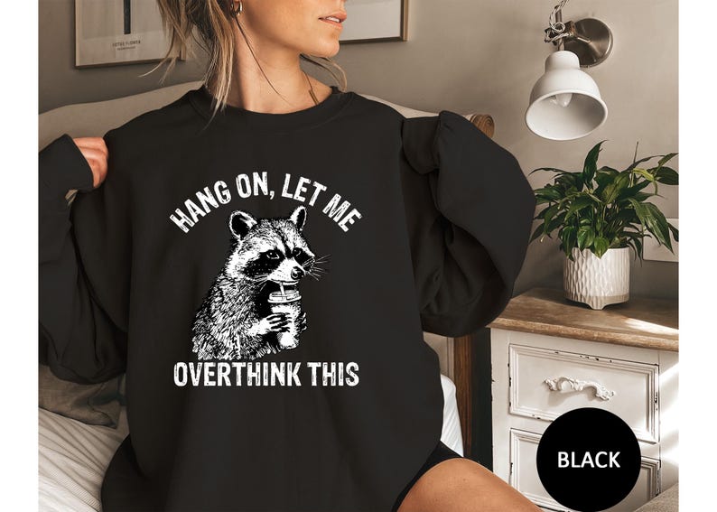 May include: Black sweatshirt featuring a raccoon holding a beverage, with the text "HANG ON, LET ME OVERTHINK THIS" in white. The raccoon graphic is detailed in black and white. The sweatshirt is a casual style.