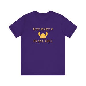 May include: Purple short-sleeved t-shirt with the text "Optimistic Since 1961" and a yellow Viking helmet graphic.