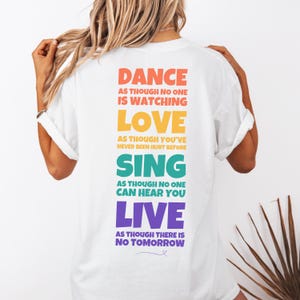 Könnte beinhalten: Weißes T-Shirt mit einem bunten Grafikdruck, auf dem steht: "Tanzen, als würde dich niemand beobachten, Lieben, als wärst du nie verletzt worden, Singen, als könnte dich niemand hören, Leben, als gäbe es kein Morgen."