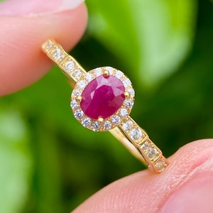 Peut inclure: Une bague en or ornée d'un grand rubis ovale, entouré d'un halo de petits diamants transparents. La monture de la bague est également ornée de petits diamants. La bague est présentée sur un fond vert flou.