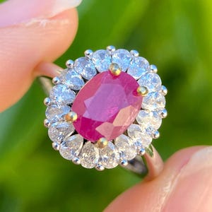 May include: A silver ring featuring a large, oval-cut ruby gemstone surrounded by a halo of smaller, clear gemstones. The ruby is a deep, vibrant red, and the surrounding stones sparkle. The ring is held against a blurred green background.