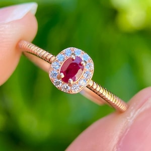 Puede incluir: Un anillo de oro rosa con una piedra central ovalada de rubí, rodeada de piedras preciosas claras más pequeñas. La banda del anillo tiene un diseño texturizado. El fondo es verde borroso.